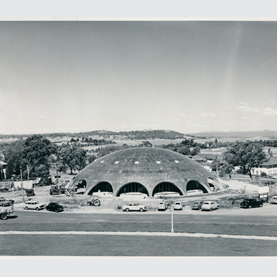 A black and white photo of Becker House on a sunny day (as it was then - now the Shine Dome), taken from across the road. 1950s era cars are parked around the dome and the vista behind the building is of low hills, paddocks and trees - very different to the Canberra of today.